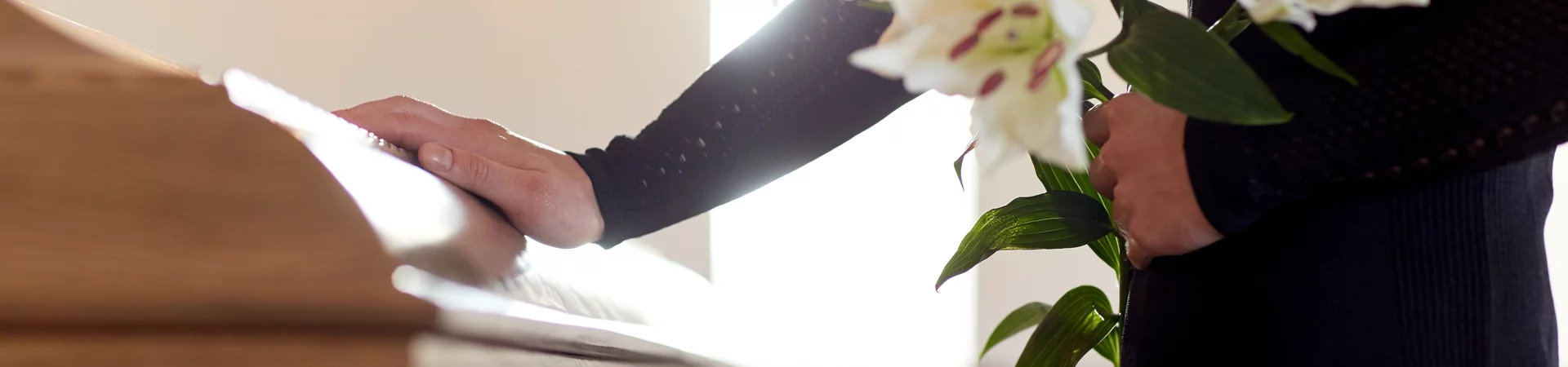 A person stands solemnly, holding a white lily before a wooden casket, representing love and tribute.